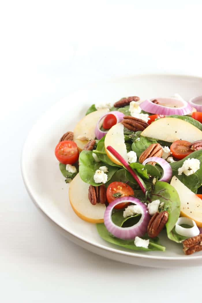 Spinach Pear Salad with Goat Cheese and Lemon Thyme Vinaigrette | There's nothing boring about this flavor-packed salad! A bed of baby spinach is topped with crisp pears, juicy tomatoes, crunchy red onions and pecans, and creamy goat cheese, and tossed in a bright and aromatic lemon thyme vinaigrette. Get the full vegetarian and gluten-free recipe via JessicaLevinson.com #SpinachSaladwithGoatCheese #EasySpinachSalad Spinach Pear Salad with Goat Cheese on White Plate